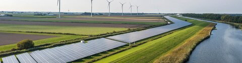 Ein Fluss, daneben PV-Anlagen, im Hintergrund Windräder