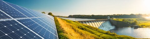 Ein Fluss, daneben PV-Anlagen, im Hintergrund Windräder