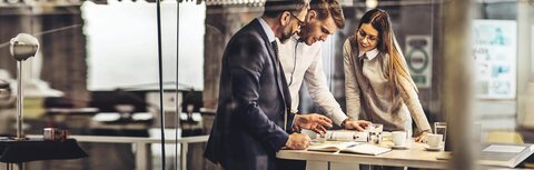 Zeitschriftenabo Drei KollegInnen, die um einen hohen Bürotisch stehen und auf eine Zeitschrift gucken