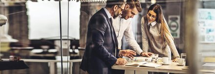 Zeitschriftenabo Drei KollegInnen, die um einen hohen Bürotisch stehen und auf eine Zeitschrift gucken