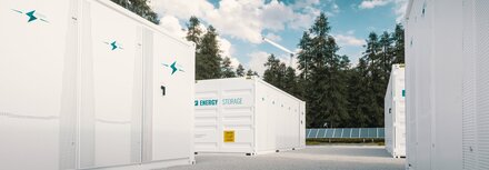 Batteriespeicher Im Vordergrund weiße Batteriespeicher, im Hintergrund Bäume, ein Windrad und eine PV-Anlage