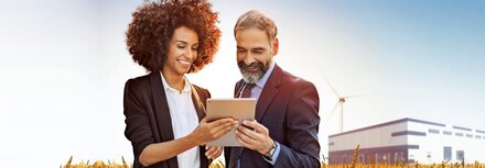 EWE business Flexload Eine Frau und ein Mann stehen in einem Getreidefeld und gucken auf ein Tablet, im Hintergrund ein Unternehmensgebäude und ein Windrad