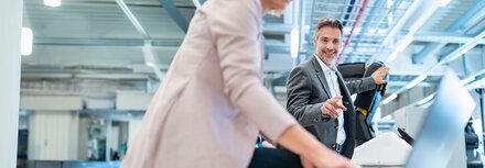 EWE business Strom fix spot Ein lächelnder Mann im grauen Anzug deutet in einer Werkhalle auf einen Laptop, den eine Frau in Benutzung hat.