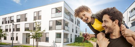 Ein weißes Mehrfamilienhaus, im Vordergrund ein Vater mit seinem Sohn auf den Schultern