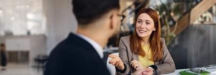 Eine Frau in Business-Kleidung erklärt einem Mann im Anzug etwas und lächelt freundlich.