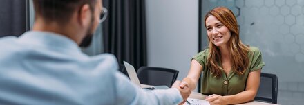 Eine lächelnde Frau schüttelt einem Mann an einem Bürotisch die Hand.