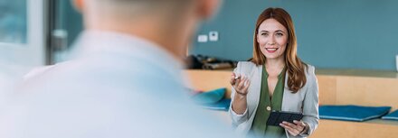 Eine Frau mit Sakko und Tablet erklärt einem Mann etwas.