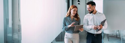 Eine Frau läuft neben einem Mann in einem Büro und erklärt ihm etwas anhand eines Tablets.