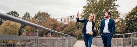 Eine Frau und ein Mann in Business-Kleidung gehen über eine Brücke, die Frau zeigt auf etwas.