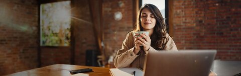 Wartung: Das System steht Ihnen schnellstmöglich wieder zur Verfügung. Junge Frau sitzt wartens vor einem Notenbook und hält eine Tasse Kafee in beiden Händen