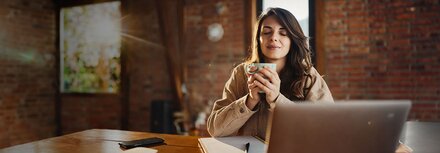 Wartung: Das System steht Ihnen schnellstmöglich wieder zur Verfügung. Junge Frau sitzt wartens vor einem Notenbook und hält eine Tasse Kafee in beiden Händen