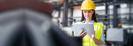Asset Tracking für Logistik und Produktion Eine Frau mit Warnweste und Helm arbeitet mit dem Tablet in einer Industriehalle.