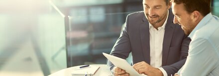 Vergleich Telekommunikationsangebote für Privat- und Geschäftskunden Ein Mann im Anzug und ein weiterer Mann in einem hellblauen Hemd sitzen an einem Tisch und schauen auf ein Tablet, vor ihnen liegen Unterlagen und Stifte.