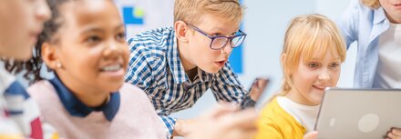 Kinder sitzen an einem Tisch in einem Klassenzimmer und schauen auf ein Tablet, dass ein Mädchen in der Hand hält.