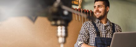Online-Marketing für kleine Unternehmen Ein Handwerker hält ein Notebook in seiner Werkstatt und lächelt.