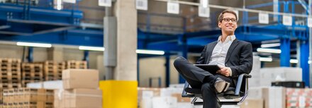 Perfekte Kombination: SD-WAN und SASE Ein Mann im Anzug sitzt auf einem Bürodrehstuhl im Lager, auf seinem Bein liegt ein Tablet.