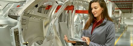 Eine Frau steht in einer Produktionshalle neben silbernen Rohlingen von Fahrzeugteilen. Sie hält in Laptop in der Hand und schaut darauf.