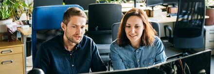 Teams-Telefonie für einen effizienten digitalen Austausch im Unternehmen Ein Mann im dunkelblauen Hemd und eine Frau im hellblauen Hemd sitzen vor zwei Bildschirmen in einem Büro.