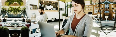 10 Energiespartipps für den Sommer Eine junge Frau arbeitet auf einer sonnenbeschienen Cafe-Terrasse unter einem Sonnenschirm an einem Laptop, im Hintergrund ein Mann