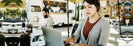 Eine junge Frau arbeitet auf einer sonnenbeschienen Cafe-Terrasse unter einem Sonnenschirm an einem Laptop, im Hintergrund ein Mann