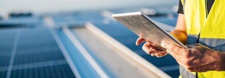 Photovoltaik Checkliste Ein Mann in gelber Warnweste hält ein Tablet in den Händen, im Hintergrund eine PV-Anlage