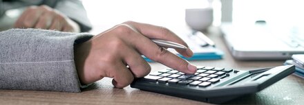 Eine Hand, die etwas auf einem Taschenrechner eingibt, der auf einem Schreibtisch liegt