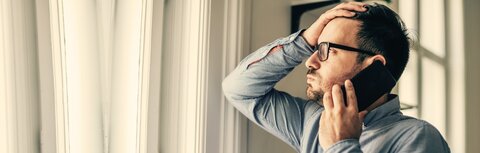 Aktuelle Trends der Cyberkriminalität Ein Mann steht im Büro, schaut durch das Fenster nach draußen, telefoniert mit einem Smartphone und langt sich mit seiner rechten Hand an den Kopf.