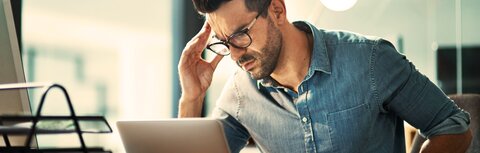 BSI-Bericht: Die Zahl der Cyberangriffe hat sich erhöht. Ein Mann sitzt am Schreibtisch vor einem Laptop, reibst sich mit seiner rechten Hand die rechte Schläfe, grübelt über einen erfolgreichen CEO-Fraud-Angriff auf sein Unternehmen.