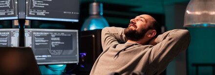 Ein Mann, der am Schreibtisch mit Bildschirmen sitzt, lächelt und die Arme hinter dem Kopf verschränkt