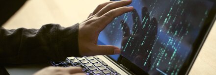 DDoS-Attacken Ein Hacker sitzt an einem Laptop und zeigt mit seiner Hand auf den Bildschirm. Auf dem Bildschirm sind Zahlen und Buchstaben zu sehen.
