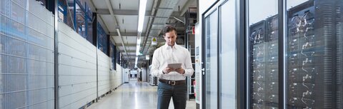 Managed Services gegen Fachkräftemangel in der IT Ein Mann im weißen Hemd steht mit einem Tablet neben einem Serverbereich in einer Fabrik.