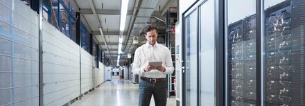 Managed Services gegen Fachkräftemangel in der IT Ein Mann im weißen Hemd steht mit einem Tablet neben einem Serverbereich in einer Fabrik.