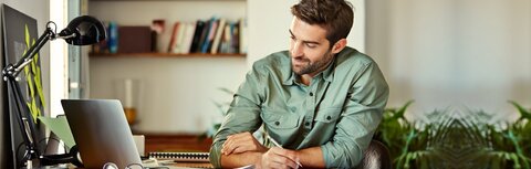 IT-Sicherheit im Homeoffice Ein Mann im grünen Hemd sitzt am Schreibtisch, guckt auf seinen Laptop-Bildschirm und macht sich dabei Notizen