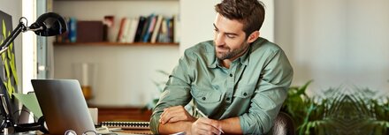 Ein Mann im grünen Hemd sitzt am Schreibtisch, guckt auf seinen Laptop-Bildschirm und macht sich dabei Notizen