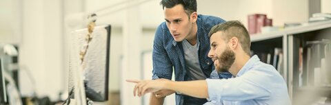 Managed Security von EWE Zwei junge Männer am Schreibtisch in einem Büro. Einer der Männer sitzt auf seinem Stuhl und zeigt mit seinem Arm auf seinen Monitor. Der andere Mann steht neben ihm und schaut auf den Monitor.