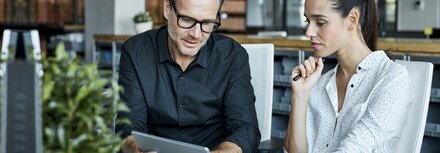 Bild: Morsa Images / DigitalVision / Getty Images Eine Frau und ein Mann sitzen an einem Schreibtisch in einem Büro und schauen sich etwas auf einem Tablet-PC an.