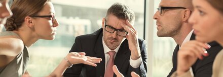 Geschäftsleute sitzen an einem Tisch. Sie diskutieren miteinander. Am Kopf des Tisches sitzt der Chef und grübelt nach.