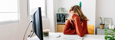 Phishing-Attacken Eine junge Frau sitzt am Schreibtisch und fasst sich nach einer erfolgreichen Phishing-Attacke mit der linken Hand an die Stirn.
