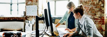 Schutz vor Ransomware Zwei Frauen stehen an einem Hochtisch und arbeiten an einem Laptop und einem Computer.