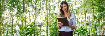 Eine Frau mit Tablet steht vor einer Reihe Pflanzen, im Hintergrund sind ein Firmengebäude und PKW zu sehen.