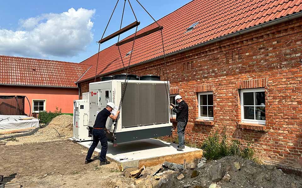 Zwei Monteure installieren eine Wärmepumpe vor einem Gebäude des Hofs Jüterborg.