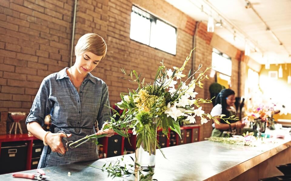 Eine Frau schneidet in einem Blumenladen eine Strauß zu, im Hintergrund eine weitere Mitarbeiterin