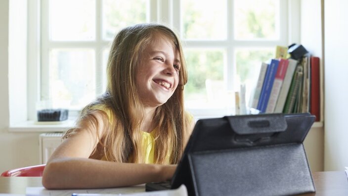 Ein Mädchen sitzt vor einem Tablet und lacht, weil es zuhause digital arbeiten kann.