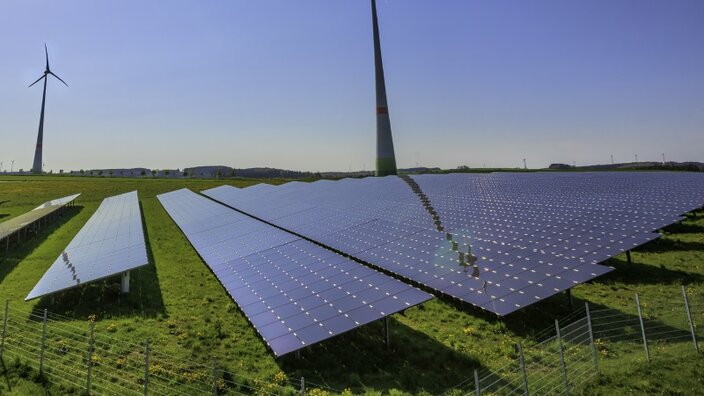 Photovoltaik-Module auf grüner Wiese, im Hintergrund Windräder