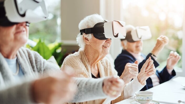 Drei Seniorinnen und Senioren sitzen an einem Tisch in einem Pflegeheim, haben Virtual Reality Brillen mit Smartphones auf und spielen gemeinsam Online-Spiele. 