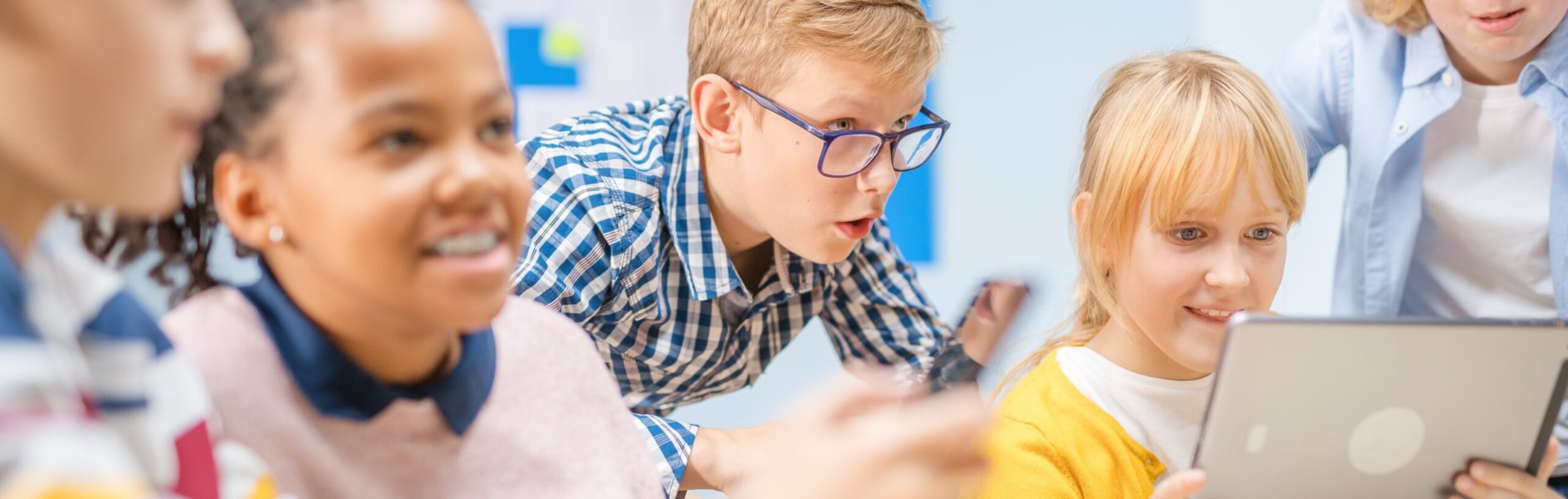Kinder sitzen an einem Tisch in einem Klassenzimmer und schauen auf ein Tablet, dass ein Mädchen in der Hand hält.