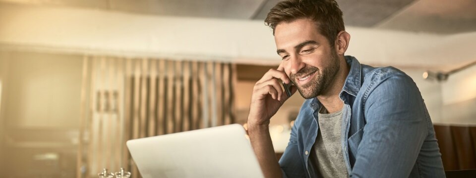 Ein Mann sitzt in einem Restaurant an einem Tisch, schaut auf den Monitor eines Laptops, lächelt und telefoniert parallel mit seinem Smartphone. 