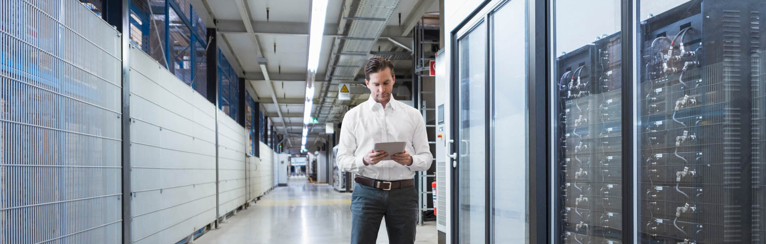 Ein Mann im weißen Hemd steht mit einem Tablet neben einem Serverbereich in einer Fabrik.
