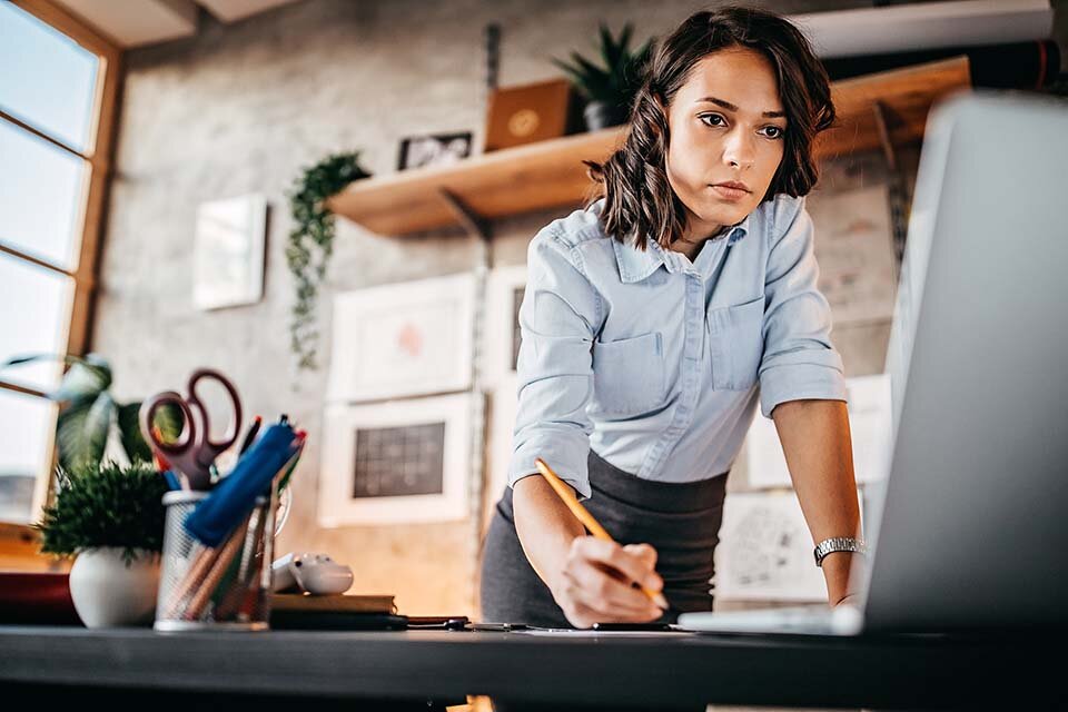 Eine Frau in Businesskleidung steht hinter einem Schreibtisch, guckt auf einen Laptop und macht sich Notizen.