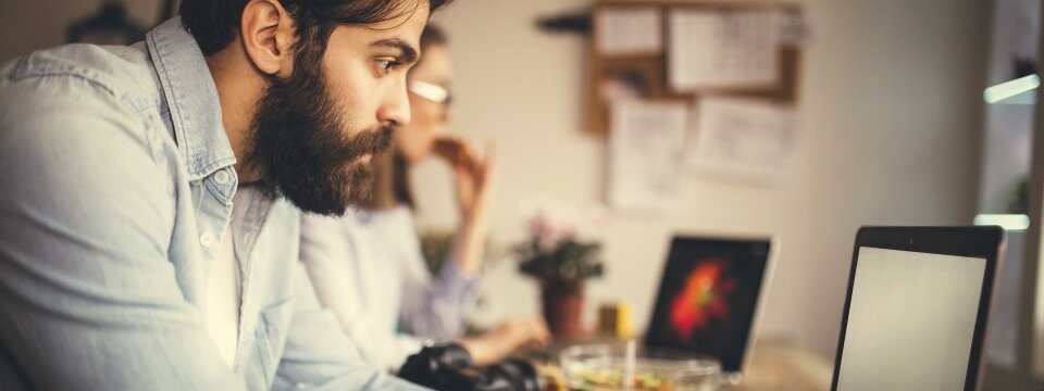 Ein Mann mit Bart sitz an einem Schreibtisch, schaut fokussiert auf sein Laptop und schreibt gerade etwas. Im Hintergrund ist eine Frau zu sehen, die ebenfalls auf ein Laptop schaut.